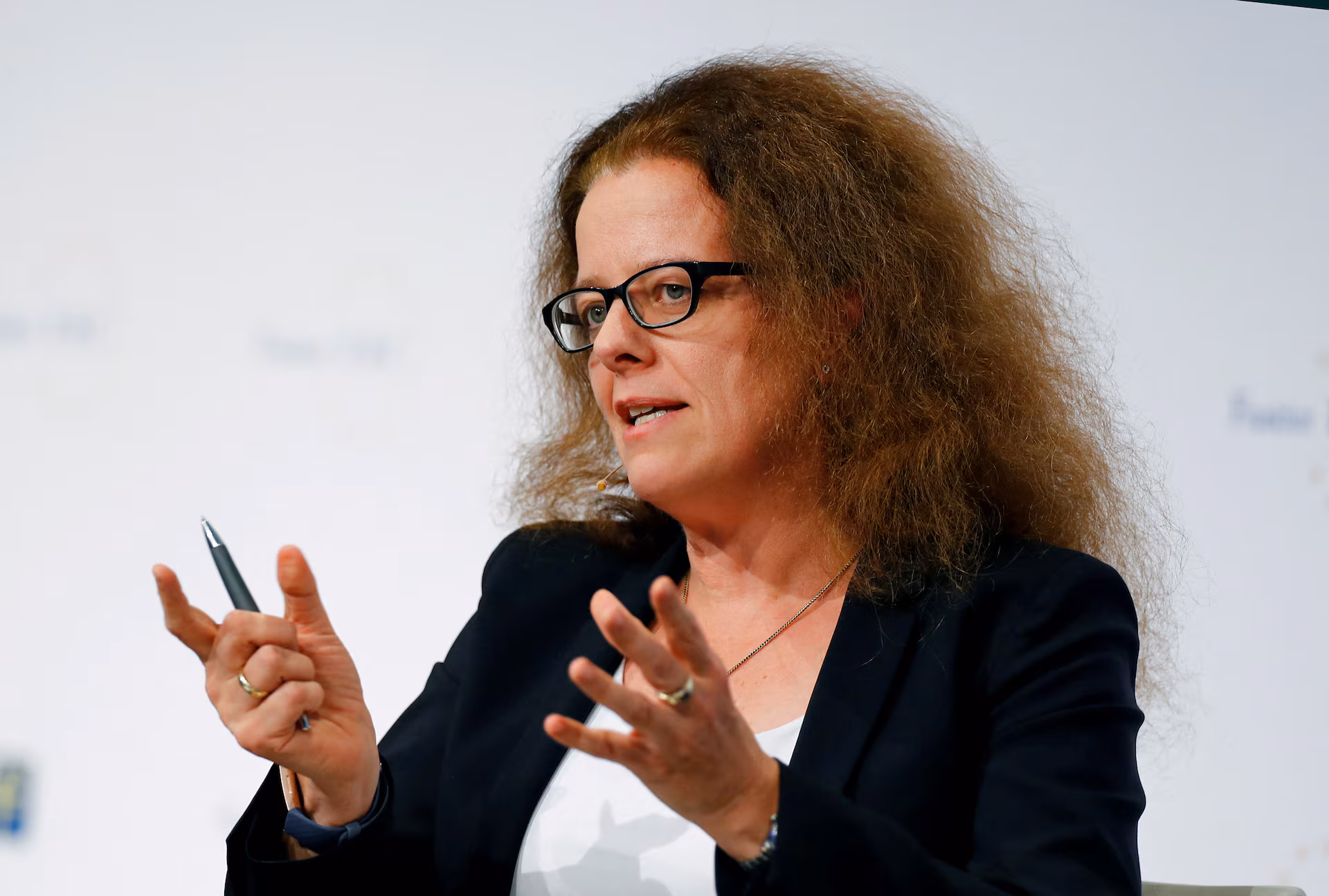 A woman with curly hair and glasses appears to be speaking or presenting, holding a pen.