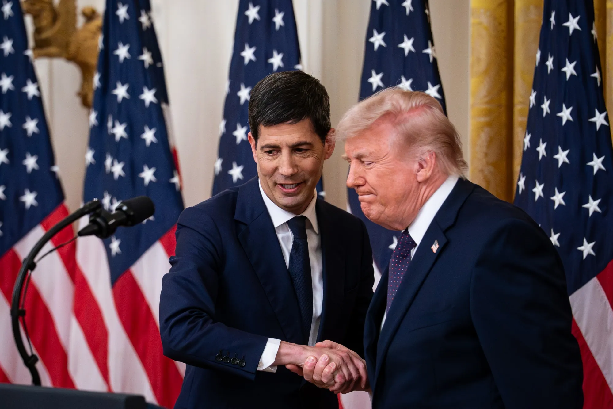 Kevin Warsh 與 Donald Trump 在一場正式活動中於美國國旗前握手。